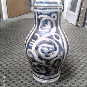 May include: A tall, bulbous ceramic beer stein featuring a dark blue and off-white glaze with intricate, swirling floral patterns. The vessel is topped with a metallic, hinged lid and has a wide, rounded body that tapers toward the neck and base.