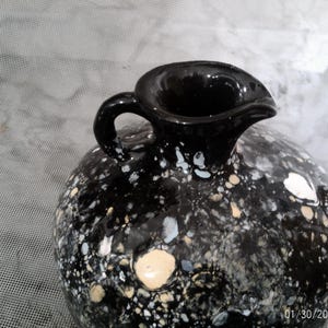 May include: Black ceramic vase with a speckled white and cream design. The vase has a wide mouth and a handle.