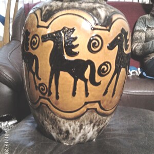 May include: A large ceramic vase featuring a tan central band decorated with black silhouettes of horses and spiral motifs. The neck and base of the vase are finished in a mottled dark brown and cream glaze, creating a distinct contrast.