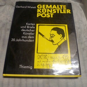 May include: A hardcover book titled Gemalte Künstler Post by Gerhard Wietek with a black cover and yellow text. The cover features a yellow square containing a black line drawing of a person smoking a pipe and handwritten text in black ink.