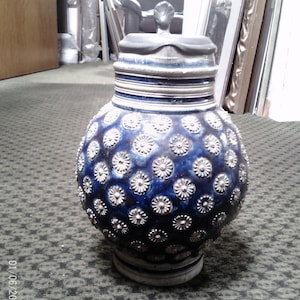 May include: A blue ceramic beer stein with a lid and handle. The body of the stein is decorated with a pattern of small, silver-colored flower-like designs. The lid and handle are dark gray.