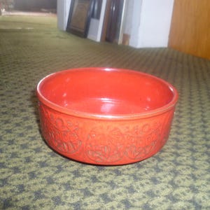Könnte beinhalten: Eine runde, rote Keramikschale mit einem dekorativen geprägten Muster auf der Außenseite. Die Schale steht auf einer gemusterten grün-schwarzen Oberfläche. Die Schale ist etwa 7.6 cm hoch und hat einen Durchmesser von 17.8 cm.