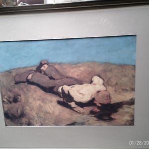 May include: A framed painting of two figures lying on a grassy field. The figures are partially obscured by the grass, but their bodies are visible. The sky is a light blue color. The painting is framed in a simple, silver frame.