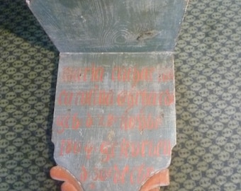 May include: A wall-mounted wooden shelf painted in a muted green with a curved top and decorative carved base. The front panel features hand-painted red lettering and the year 1818. The wood grain is visible through the weathered green paint finish.
