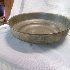 May include: A large, round, metal serving bowl with two handles. The bowl is made of a dull, aged metal and has a slightly textured surface.