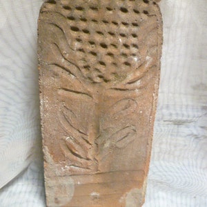 May include: A terracotta tile with a raised design of a face with closed eyes and a stylized plant growing from the forehead.