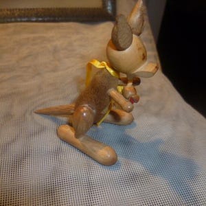 May include: A wooden kangaroo figurine with a yellow ribbon around its neck. The kangaroo is holding a small wooden joey in its pouch.