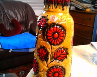 May include: A tall, ceramic vase with a textured, tan surface. The vase features red and black floral designs and a dark rim. The vase is approximately 12 inches tall and has a wide base.