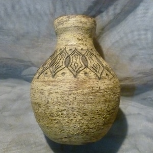 Pode incluir: Vaso de cerâmica bege com superfície texturizada e um padrão geométrico preto decorativo ao redor do pescoço. O vaso tem um corpo arredondado e uma abertura estreita, adequado para exibir flores ou como objeto decorativo.