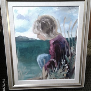 May include: Oil painting of a person in a field, framed in a white and gold frame. The person is wearing a purple shirt and blue pants, with blonde hair. The background features a green field, blue sky, and mountains.
