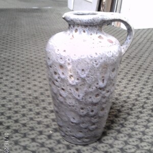 May include: A tall, ceramic vase with a handle, featuring a textured, speckled design in shades of white, gray, and brown. The vase has a wide opening and a curved handle, suitable for decorative purposes.