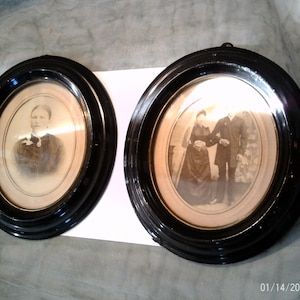 Può includere: Due ritratti antichi ovali incorniciati. Le cornici sono nere con finitura lucida. I ritratti sono in bianco e nero, raffiguranti persone in abiti formali. Un ritratto mostra una singola persona, l'altro ne mostra tre.