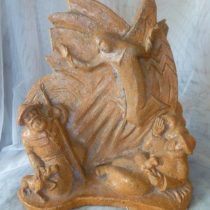 May include: A tan-coloured relief sculpture depicting a central figure with wings hovering above two kneeling figures, one holding a staff and accompanied by a small animal, while the other figure sits with hands raised, all rendered in a textured material.