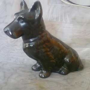 Può includere: Una statuetta di cane in ceramica con una glassa verde scuro e marrone. Il cane è seduto con la testa girata di lato.