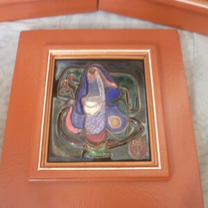 May include: A square ceramic tile featuring a central abstract figure in shades of blue, green, and purple, set within a thick, matte terracotta-colored frame. The tile has a glossy finish and includes a small circular stamp with a symbol in the corner.