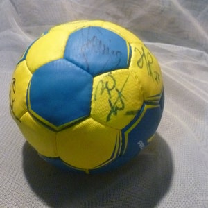 Puede incluir: Un balón de balonmano amarillo y azul con costuras negras y varias firmas en marcador negro.