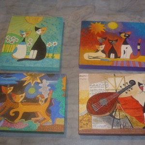May include: Four square paintings featuring stylized cat illustrations. Each painting has a different background and cat arrangement. Colors include blue, orange, yellow, black, and white. One painting shows a cat with a musical instrument.