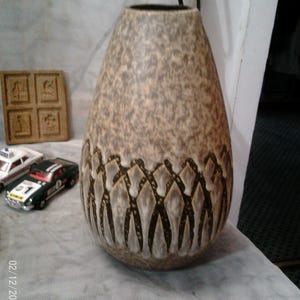 May include: A tall, teardrop-shaped vase with a textured, speckled brown and tan surface. The base features a repeating pattern of stylized figures in a darker brown. The vase has a dark brown interior and a wide opening.