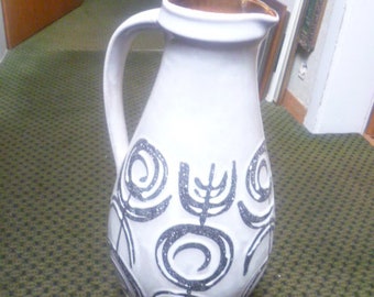 May include: A white ceramic pitcher with a brown rim and a black and white geometric design. The pitcher has a wide base and a narrow neck.