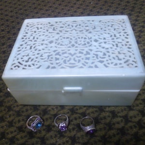 May include: A white jewelry box with a decorative, openwork lid and three rings with colorful gemstones. The rings are silver-toned with blue, purple, and violet stones. The box is rectangular and closed.