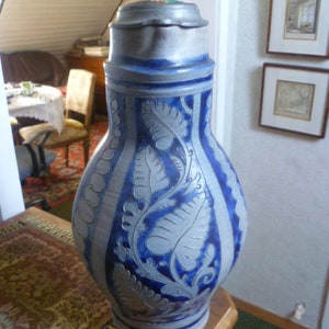 May include: A tall, bulbous stoneware beer stein featuring a grey and cobalt blue salt-glazed finish. The vessel is decorated with embossed fern leaf patterns and vertical stripes, topped with a hinged pewter lid with a thumb lift mechanism.