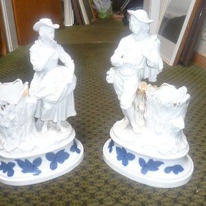 Peut inclure: Deux figurines en porcelaine blanche représentant une personne portant un chapeau appuyée contre un vase en forme de souche d'arbre. Les bases ovales sont ornées de motifs de feuilles bleues et les souches ont des accents dorés.