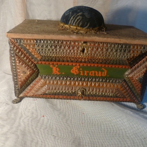 Op de afbeelding: Een houten doos met een scharnierend deksel en een kleine lade. De doos is versierd met ingewikkelde geometrische patronen in tinten groen, oranje en bruin. De doos heeft een klein metalen handvat en een naaldenkussen bovenop. De doos is gelabeld "K. Giraud".