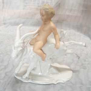 Könnte beinhalten: Eine Porzellanfigur, die ein nacktes Kind mit hellem Haar auf einer weißen Ziege reitend darstellt. Die Ziege hat geschwungene Hörner und ist in einer dynamischen Pose eingefangen. Das gesamte Stück hat eine glänzende Oberfläche und ist auf einem ovalen Sockel montiert, was die detaillierte Handwerkskunst der skulptierten Formen von Kind und Tier unterstreicht.
