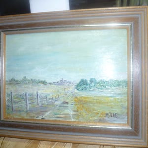 May include: A framed oil painting depicting a rural landscape with a dirt road leading to a small town in the distance. The painting is done in muted tones of blue, green, and brown, and features a fence along the side of the road. The painting is signed "M.R. 1935" in the lower right corner.