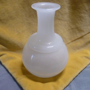 Puede incluir: Un jarrón de vidrio blanco opaco con base redondeada y cuello delgado. El jarrón tiene un acabado liso y brillante y un borde ancho y acampanado. El jarrón está sobre una superficie amarilla.