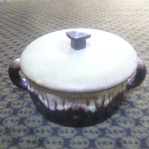 Op de afbeelding: Een keramische braadpan met deksel, een crèmekleurig deksel en een donkerbruin handvat aan elke kant. De schaal heeft een tweekleurig glazuur, met een lichtgroene bovenkant en een donkerbruine onderkant.