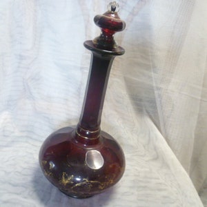 Può includere: Una caraffa di vetro rosso scuro con un design dipinto in oro e un tappo in vetro trasparente. La caraffa ha un corpo rotondo e un collo lungo e sottile.