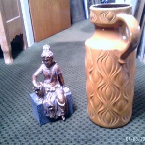 May include: A brown ceramic vase with a handle and a textured, repeating pattern. Beside it is a small bronze-colored statue of a seated figure on a blue block. The items are on a patterned green surface.