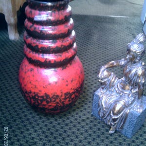 Puede incluir: Un jarrón grande de cerámica roja y negra con una superficie texturizada y un interior negro. Junto a él, una estatua de una figura sentada con detalles dorados, sobre una base de piedra azul. El jarrón tiene una base bulbosa y una sección superior cilíndrica apilada.