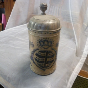 May include: A cylindrical stoneware beer stein with a tan body and blue cobalt decorative patterns featuring a central crest and floral motifs. The vessel is topped with a fitted pewter lid that includes a small, rounded finial handle.