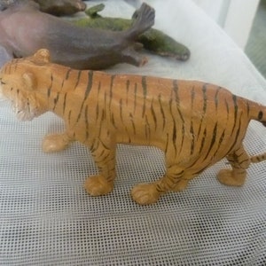 Könnte beinhalten: Eine Spielzeug-Tigerfigur mit orangefarbenen und schwarzen Streifen, die auf einer weißen Oberfläche steht. Der Tiger ist eine detaillierte Miniatur, die die Merkmale des Tieres zeigt. Andere Tierfiguren sind im Hintergrund zu sehen.