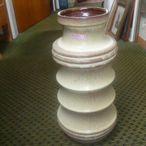 May include: A tall, cream-coloured ceramic vase with a brown rim and a textured surface. The vase has a series of ridges that create a unique, layered look. The vase is labelled "Scheurich" in pink.