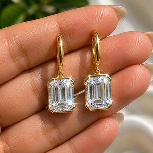 May include: A pair of gold hoop earrings featuring rectangular emerald-cut clear stones suspended from each hoop. The stones are set in polished gold frames, reflecting light with a faceted surface. The design consists of a simple circular hoop connected to a dangling, geometric, clear gemstone pendant.