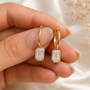 May include: A pair of gold hoop earrings featuring emerald-cut clear stones in bezel settings. The earrings consist of a polished gold hoop attached to a rectangular, faceted white gemstone, creating a drop design with a metallic finish.