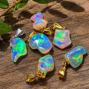Op de afbeelding: Zes onregelmatig gevormde opaalhangers met levendige flitsen van blauw, groen en oranje kleurenspel. Vijf hangers hebben goudkleurige metalen oogjes, terwijl één hanger een zilverkleurig metalen oogje heeft, getoond op een gestructureerd houten oppervlak.