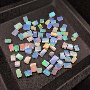 Puede incluir: Una colección de pequeñas piedras de ópalo iridiscente en varios tonos de blanco, azul y verde. Las piedras son de forma rectangular y tienen un acabado liso y pulido.