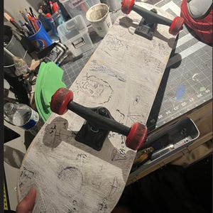May include: A skateboard with a white deck featuring hand-drawn black ink sketches and mathematical equations. The skateboard has black metal trucks and four red wheels. The deck surface shows visible scratch marks and various doodles.