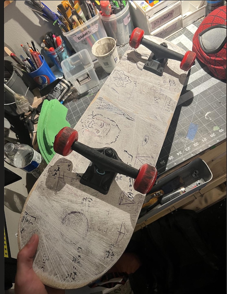 May include: A skateboard with a white deck featuring hand-drawn black ink sketches and mathematical equations. The skateboard has black metal trucks and four red wheels. The deck surface shows visible scratch marks and various doodles.