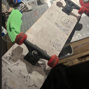 May include: A skateboard with a light-colored deck featuring various black ink drawings, including a portrait, geometric shapes, and mathematical equations. The skateboard has two black metal trucks and four red wheels, showing signs of wear.