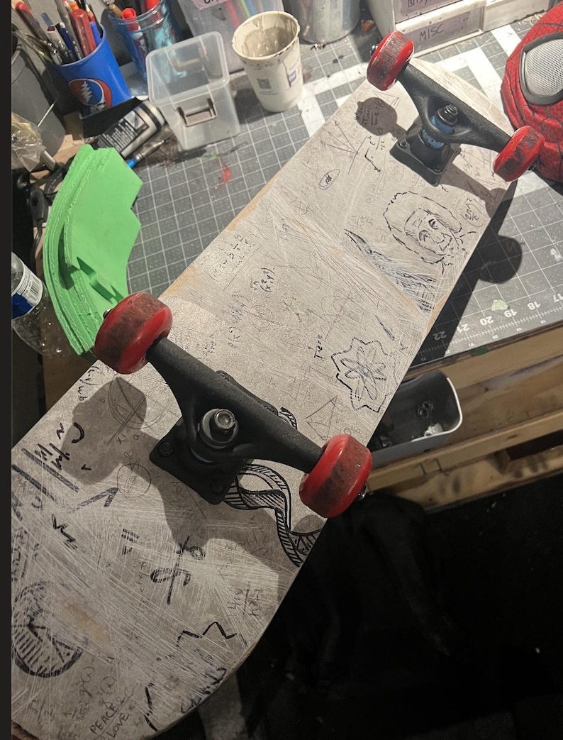May include: A skateboard with a light-colored deck featuring various black ink drawings, including a portrait, geometric shapes, and mathematical equations. The skateboard has two black metal trucks and four red wheels, showing signs of wear.