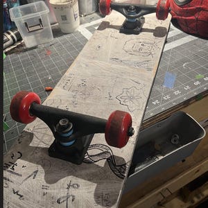 May include: A skateboard with a light gray deck featuring black hand-drawn illustrations of mathematical equations, geometric shapes, and a portrait. The skateboard has black metal trucks, light blue bushings, and four red wheels.
