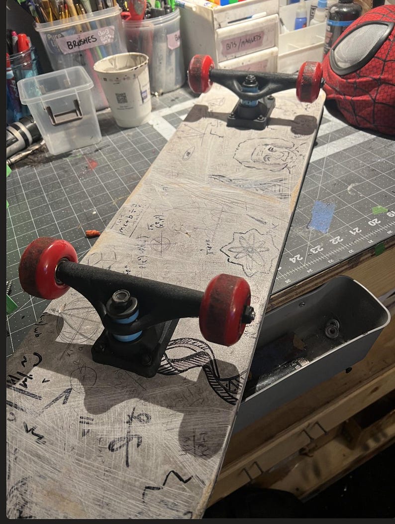 May include: A skateboard with a light gray deck featuring black hand-drawn illustrations of mathematical equations, geometric shapes, and a portrait. The skateboard has black metal trucks, light blue bushings, and four red wheels.