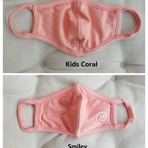 May include: Two coral-colored face masks for kids. The top mask is plain, while the bottom mask has a white smiley face embroidered on it. Both masks have elastic ear loops.