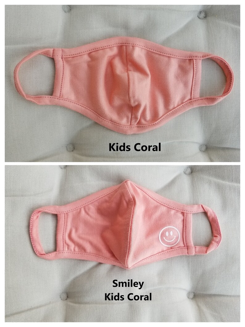 May include: Two coral-colored face masks for kids. The top mask is plain, while the bottom mask has a white smiley face embroidered on it. Both masks have elastic ear loops.