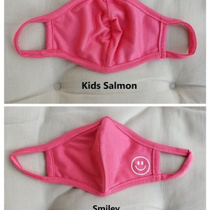 May include: Pink kids face mask with a white smiley face design. The mask is made of a soft, breathable fabric and has adjustable ear loops.